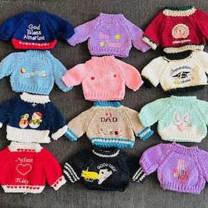 May include: Twelve knitted sweaters with various designs and colors. The sweaters have embroidered designs such as "God Bless America", "My First Teddy", "Good Luck", "Happy Birthday", "Dad", "Congratulations Graduate", "Gobble Gobble", and more.
