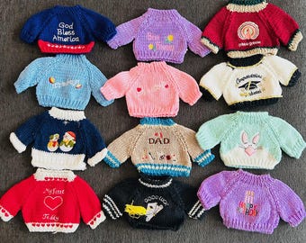 May include: Twelve knitted sweaters with various designs and colors. The sweaters have embroidered designs such as "God Bless America", "My First Teddy", "Good Luck", "Happy Birthday", "Dad", "Congratulations Graduate", "Gobble Gobble", and more.