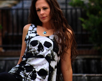 May include: A woman with long brown hair is wearing a black and white tank top with a skull print. She is wearing a silver necklace with a blue gemstone pendant. The background is a blurry image of a fountain.