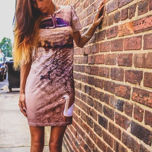 May include: A woman wearing a brown and white dress with a swan design. The dress has a short sleeve and a fitted silhouette. The woman is standing in front of a brick wall.