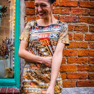 Può includere: Una donna indossa un vestito colorato con motivi e dettagli dorati. È in piedi davanti a un muro di mattoni e sorride.