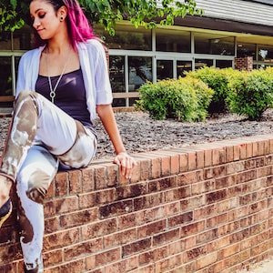 Può includere: Una donna con i capelli rosa è seduta su un muro di mattoni indossando un cardigan bianco, una canotta nera e leggings mimetici. Indossa tacchi alti neri con dettagli dorati.