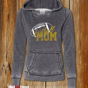 May include: A gray hooded sweatshirt with a distressed look. The front of the sweatshirt features a gold glitter design of a football with the words "football mom" in gold glitter.