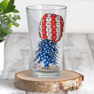 May include: A clear pint glass featuring a patriotic pineapple design. The pineapple is decorated with red and white stripes and blue stars, resembling the American flag. The glass sits on a wooden coaster.