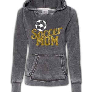 May include: A gray hoodie with a distressed look. The hoodie has a large gold glitter graphic that says "Soccer Mom" with a white soccer ball above the text.