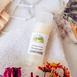 May include: A white deodorant stick with a green and white label that reads "Natural Sounds Farm Aluminum Free Deodorant Clean Cotton". The stick is on a white towel with orange and pink flowers.