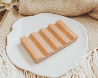 May include: A wooden soap dish with five ridges on a white plate. The dish is made of light brown wood and has a natural finish.