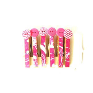 May include: Six pink wooden clothespins with decorative patterns and button accents. The clothespins have a pink and white design with various patterns, including chevrons, polka dots, and floral motifs. The buttons are pink and add a touch of whimsy.