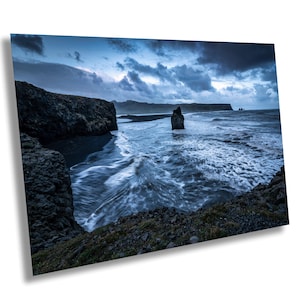 May include: A landscape photograph of a black sand beach with waves breaking on the shore. Dark, rocky cliffs border the scene, and a large rock formation is in the water. The sky is filled with dramatic, dark clouds.