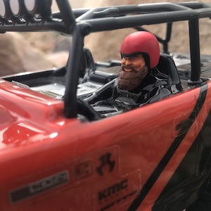 Puede incluir: Un jeep rojo de juguete con una jaula de seguridad negra y una figura de conductor negra y roja que lleva un casco rojo. El jeep tiene una franja negra y blanca en el lateral y una pegatina KMC.