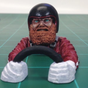 Könnte beinhalten: Miniaturfigur eines Rennfahrers mit schwarzem Helm und rotem Rennanzug. Der Fahrer hat einen detaillierten, strukturierten orangefarbenen Bart und intensive blaue Augen, die ein schwarzes Lenkrad umklammern. Die Handschuhe der Figur sind weiß.