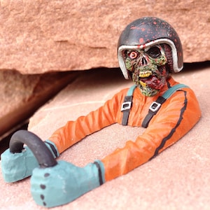Pode incluir: Uma estatueta de zumbi com um macacão de corrida laranja e um capacete preto com respingos vermelhos. O zumbi tem pele verde e vermelha e segura um volante preto com luvas azuis. A estatueta está em um fundo de pedra marrom-avermelhada.