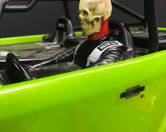 May include: A green toy car with a skeleton figure in the driver's seat. The skeleton is wearing a black racing suit with a red and white Sparco logo. The car has a black roll cage and a black door handle.