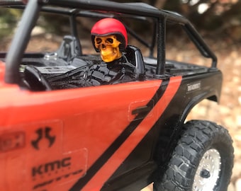 May include: A red and black toy truck with a skull figure wearing a red helmet in the driver's seat. The truck has a black roll cage and a black and red stripe design. The truck has the words "KMC" and "DEADBOLT" on the side.