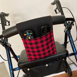 May include: A black walker with a red and black plaid fabric pouch attached to the handlebars. The pouch has two pockets, one holding a remote control and the other holding a cell phone.
