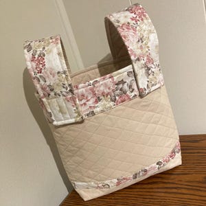 May include: A quilted fabric tote bag featuring a solid beige diamond-stitched body and floral patterned fabric accents in shades of pink, cream, and brown on the top border and the two sturdy vertical handles attached to the bag.