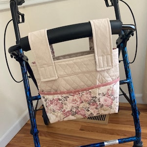 May include: A beige quilted walker bag with a floral print pocket. The bag is attached to a blue and black walker. The floral print features pink and white roses.