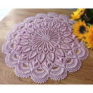 May include: A delicate, crocheted doily in a soft lavender color. The doily features a circular design with intricate details and a scalloped edge.