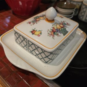 May include: A white ceramic butter dish with a lid decorated with a fruit basket design. The lid has a white knob and is decorated with colorful fruits. The dish has a green lattice pattern and a yellow trim. The dish sits on a matching white tray.