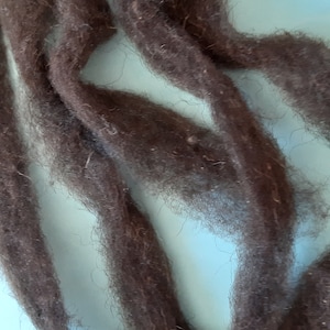 Könnte beinhalten: Dunkelbraune Wollroving, eine Naturfaser, die zum Spinnen, Filzen und für andere Handarbeiten verwendet wird. Die Wolle ist weich und flauschig, mit einem leicht strukturierten Aussehen. Das Roving wird vor einem hellblauen Hintergrund gezeigt.