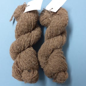 Könnte beinhalten: Zwei Stränge brauner Wolle mit weißen Anhängern. Das Garn ist fest verdreht und zeigt eine strukturierte Oberfläche. Der Hintergrund ist einfarbig hellblau.
