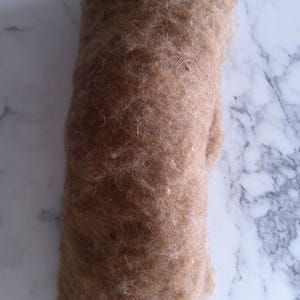Könnte beinhalten: Ein gerolltes Bündel hellbrauner Fasern, wahrscheinlich Wolle oder ein ähnliches Material, wird vor einem marmorierten Hintergrund gezeigt. Die Fasern wirken weich und strukturiert, mit einem natürlichen, erdigen Ton. Das Bündel ist dicht gepackt, was darauf hindeutet, dass es zum Basteln bereit ist.