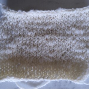May include: A close-up of a white knitted fabric swatch. The fabric has a textured, bumpy surface and a slightly uneven edge.