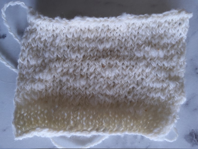 May include: A close-up of a white knitted fabric swatch. The fabric has a textured, bumpy surface and a slightly uneven edge.