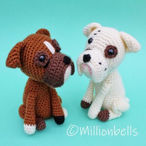 May include: Two crocheted boxer dog plushies, one brown and white, the other white with brown spots. Both are sitting and looking at each other. The background is a light turquoise.
