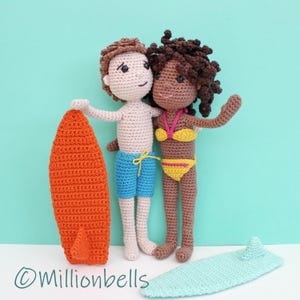 Könnte beinhalten: Zwei gehäkelte Puppen, eine mit brauner Haut und eine mit weißer Haut, stehen zusammen und halten ein leuchtend orangefarbenes Surfbrett. Die Puppe mit brauner Haut trägt einen gelben Bikini und die Puppe mit weißer Haut trägt blaue Badehosen. Die Puppen stehen auf einem hellblauen Surfbrett.