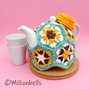 Op de afbeelding: Een turquoise gehaakte theepotwarmer met gele, witte en bruine bloemmotieven. De warmer zit op een witte theepot met een kurken onderzetter eronder. Twee witte theekopjes staan op de achtergrond.