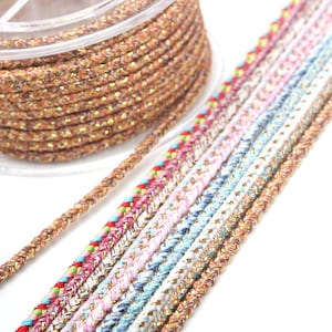 Puede incluir: Un carrete de cordón trenzado dorado y marrón con una variedad de cordones trenzados de colores en el fondo.