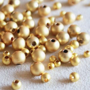 Könnte beinhalten: Eine Sammlung goldfarbener Perlen in verschiedenen Größen. Die Perlen sind rund mit mattem Finish und einem Loch in der Mitte. Sie sind auf einer hellen Oberfläche verstreut und bilden eine strukturierte und optisch ansprechende Anordnung.