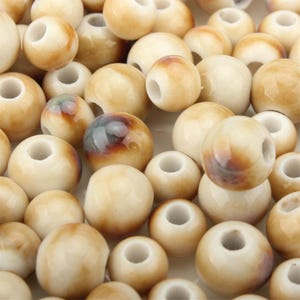 May include: A close-up of a pile of beige beads with a glossy finish. The beads are round with a hole through the centre. Some beads have brown and grey markings. Ideal for jewellery making and craft projects.