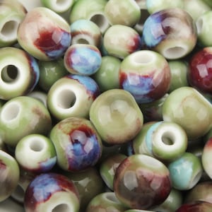 Könnte beinhalten: Nahaufnahme einer Sammlung von Keramikperlen in verschiedenen Größen und Farben. Die Perlen haben eine glänzende Oberfläche und weisen eine Mischung aus grünen, roten, blauen und cremefarbenen Farbtönen auf. Jede Perle hat ein Loch in der Mitte.