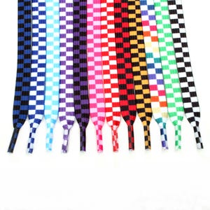 May include: A collection of twelve flat shoelaces arranged side by side, each featuring a distinct two-tone checkered pattern. The colors include black, blue, light blue, purple, pink, red, orange, yellow, green, and white in various combinations.