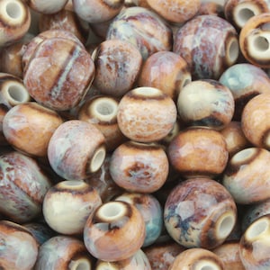 May include: A collection of round ceramic beads featuring a glossy finish with a marbled pattern in shades of brown, tan, cream, and hints of blue. Each bead has a smooth, spherical shape with a central hole and a distinct dark brown ring.