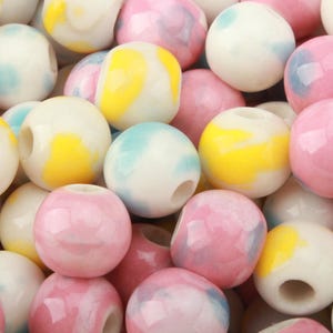 May include: A collection of round, smooth beads featuring a glossy finish. The beads are patterned with marbled swirls of pastel pink, soft yellow, and light blue colours against a white base, each containing a central hole for threading purposes.