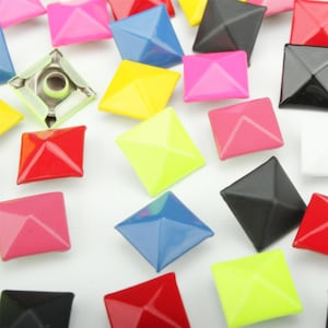 May include: A collection of square pyramid-shaped studs in various colors including red, blue, yellow, black, white, and pink. Each stud features a smooth, glossy finish with a pointed top and a metal prong attachment visible on the underside.