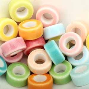 Könnte beinhalten: Eine Ansammlung von bunten Keramikperlen in Donutform, in verschiedenen Pastelltönen wie Gelb, Rosa, Orange, Grün und Blau. Die Perlen haben eine glänzende Oberfläche und ein Loch in der Mitte.