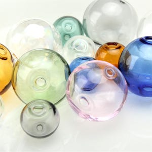 Puede incluir: Una colección de esferas de vidrio transparentes y coloridas de varios tamaños. Las esferas son en tonos de azul, verde, rosa, naranja y transparente. Cada esfera tiene un agujero. Los objetos están dispuestos cerca, creando una exhibición visualmente atractiva.