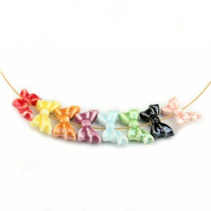 May include: A strand of colorful ceramic bow-shaped beads in a rainbow of colors, including red, yellow, orange, purple, light blue, green, black, and pink. The beads are strung on a thin gold wire against a white background.