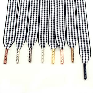 Peut inclure: Huit lacets plats avec un motif à carreaux vichy noir et blanc. Chaque lacet est terminé par un embout métallique distinct, incluant des finitions cuivre, argent, or, or rose, bronze et noir, disposés en une rangée verticale parallèle.