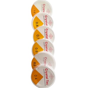 May include: Six white round containers with yellow and red text. The containers are stacked on top of each other. The text on the containers reads "Crystal Tec" and "Crystal" with different sizes listed: 1.0, 0.8, 0.7, 0.6, 0.5, and 0.4.