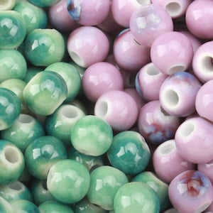 May include: A collection of round ceramic beads featuring a glossy finish. The beads are coloured in shades of light green and soft lavender, with some showing subtle mottled blue and purple accents. Each bead has a central hole for threading.