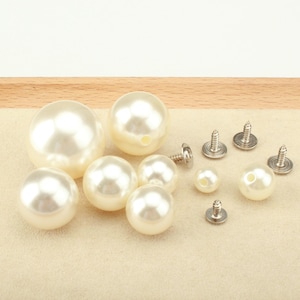 May include: A collection of off-white spherical faux pearl beads in various sizes, some featuring central holes, alongside several silver-toned metal screw-back fasteners designed for attaching the beads to fabric or other materials.