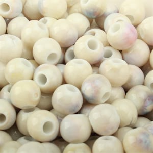 Könnte beinhalten: Eine Ansammlung runder, cremeweißer Perlen mit glatter, glänzender Oberfläche. Jede Perle hat ein mittiges Loch und weist dezente, zarte Wirbel in Hellrosa und Blau auf, die ein sanftes, marmoriertes Erscheinungsbild auf dem Haufen erzeugen.