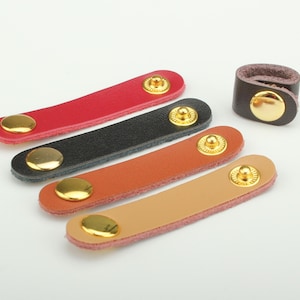 Op de afbeelding: Een verzameling leren kabel organizers in rood, zwart, oranje en beige, elk met een gouden drukknoopsluiting. Een bruine leren lus met een gouden drukknoop is ook inbegrepen. Voor kabelbeheer.