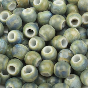 Puede incluir: Una colección de cuentas de cerámica redondas con un esmalte brillante y moteado en tonos verde salvia, azul apagado y crema. Cada cuenta tiene una superficie lisa y redondeada con un orificio central de acabado interior blanco limpio.