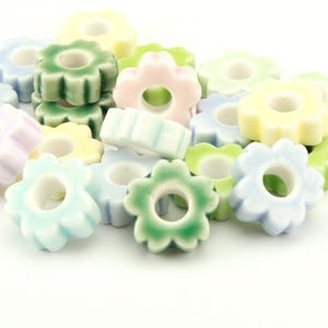 Könnte beinhalten: Eine Ansammlung kleiner, blumenförmiger Perlen in verschiedenen Pastellfarben wie Hellgrün, Blassgelb, Zartrosa und Hellblau. Jede Perle hat eine glatte, glasierte Oberfläche und ein Loch in der Mitte und ist in einem losen Haufen angeordnet.